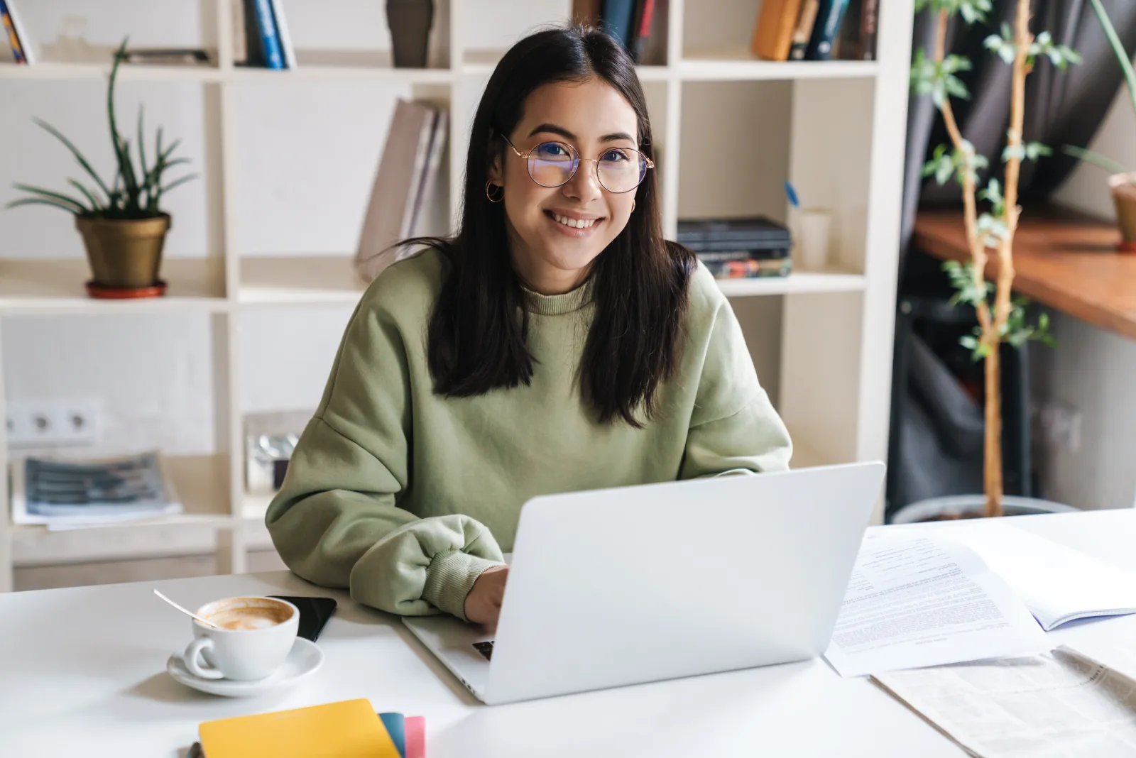 A woman in a green sweater working at a laptop at a tidy home desk with a coffee cup.