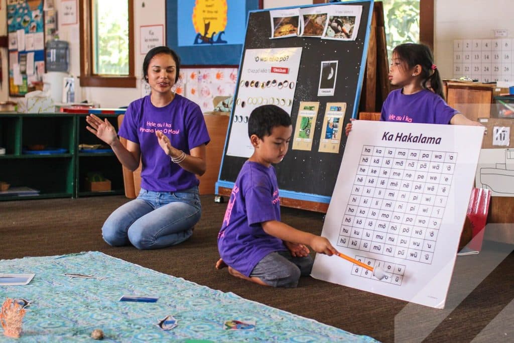 A classroom in which the Hawaiian language is being taught, with a teacher and a two children holding an alphabet chart