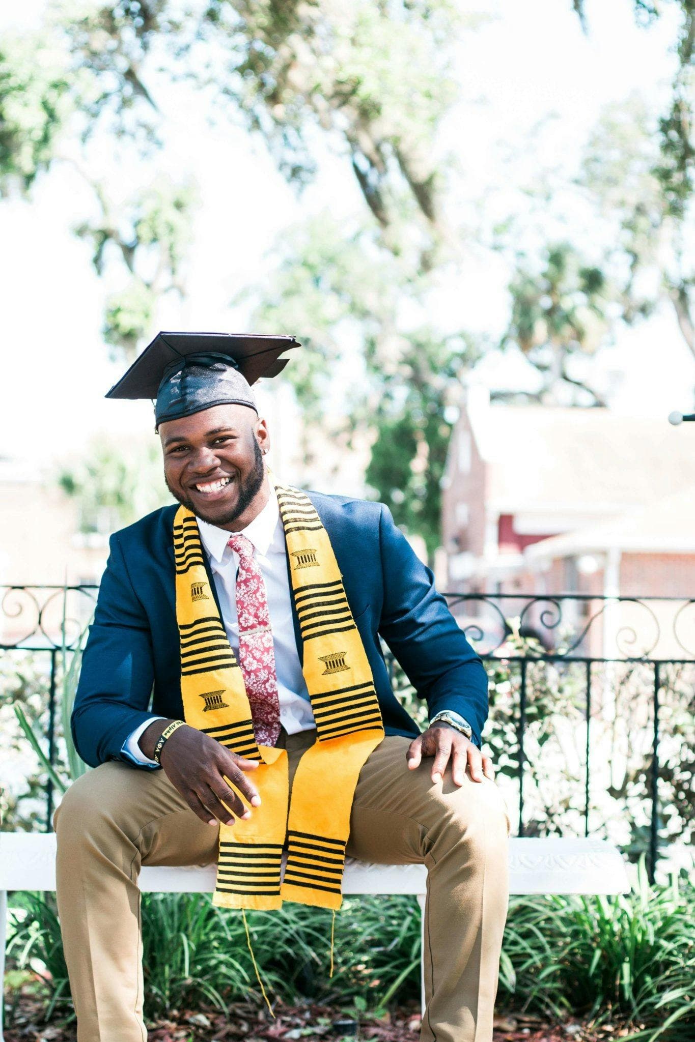 A photo of a man in a graduation cap, a suit jacket, and a yellow scarf, sitting outside in a garden.