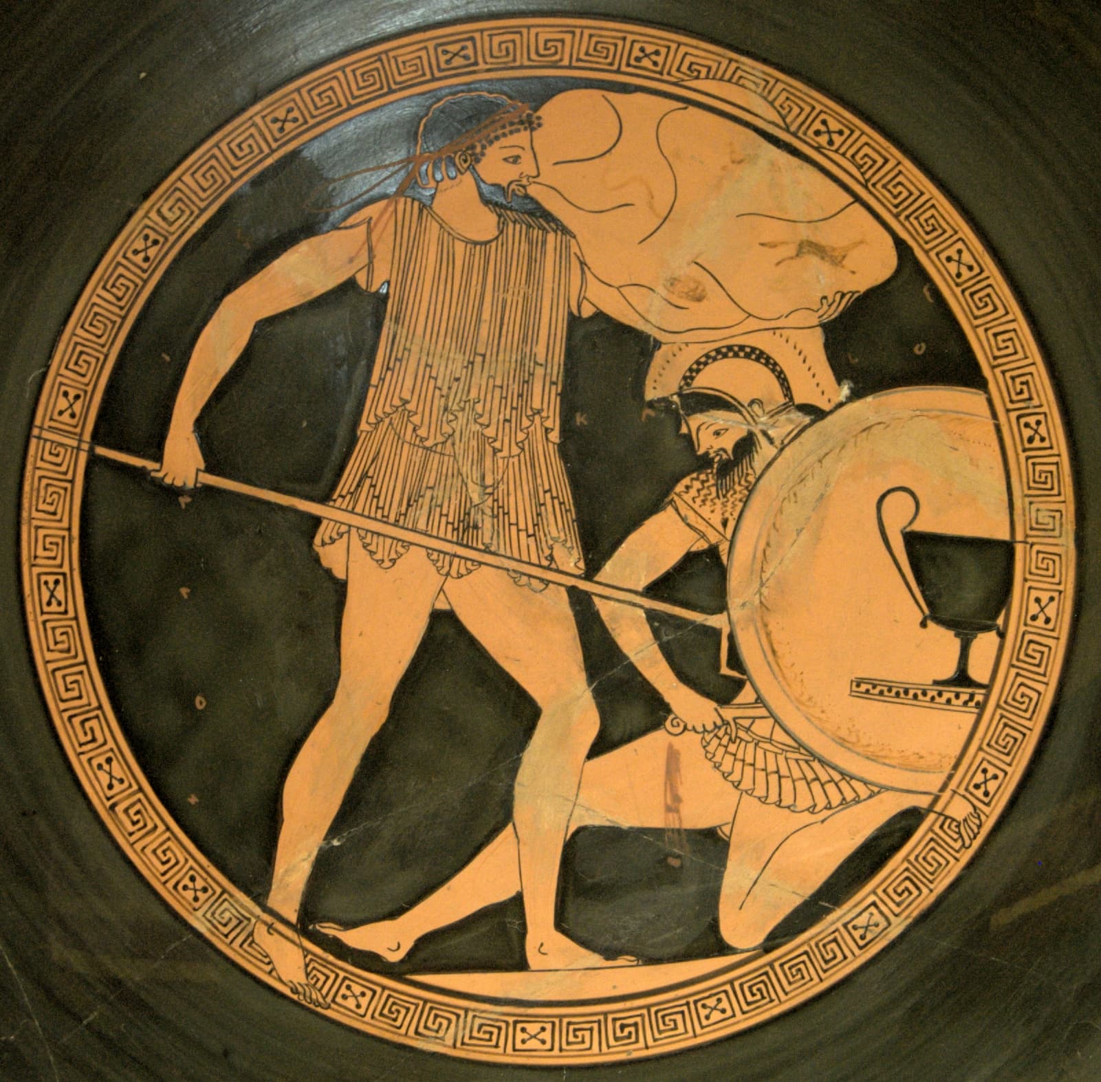 A piece of Ancient Greek pottery shows a standing man with a beard stabbing a man on his knees in his stomach with a spear. The man on the ground is , dressed as a hoplite and holds a large shield.