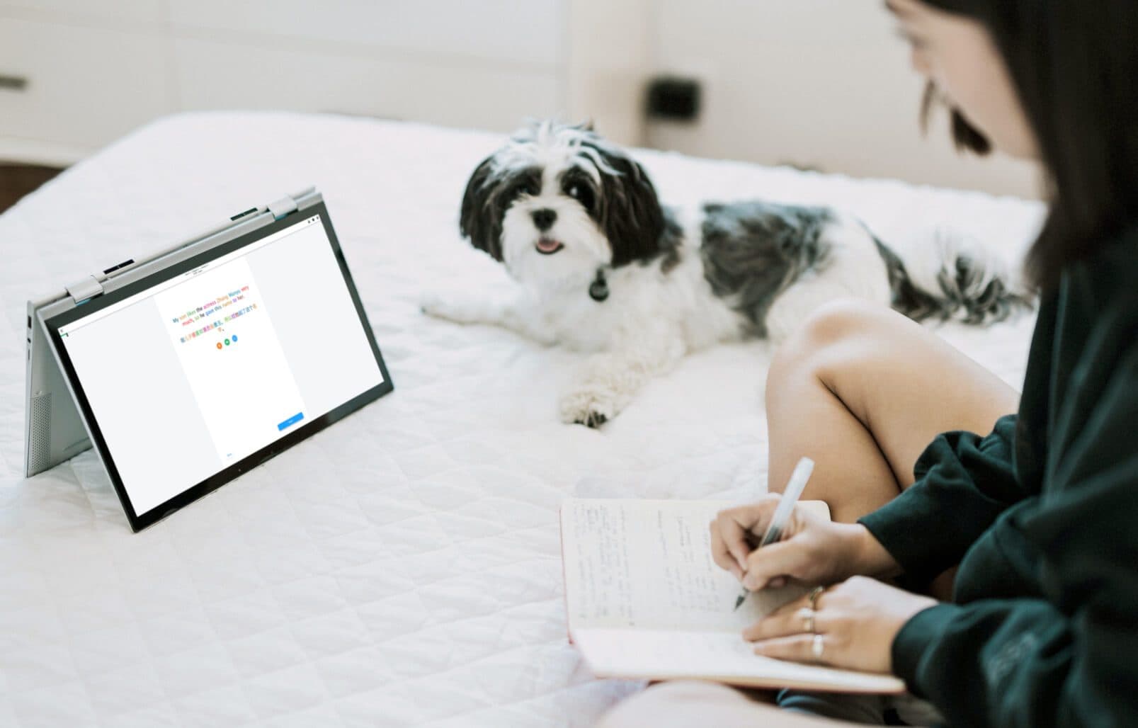 A woman sits on her bed with a small dog, writing in a notebook while she uses the Mango app on an ipad