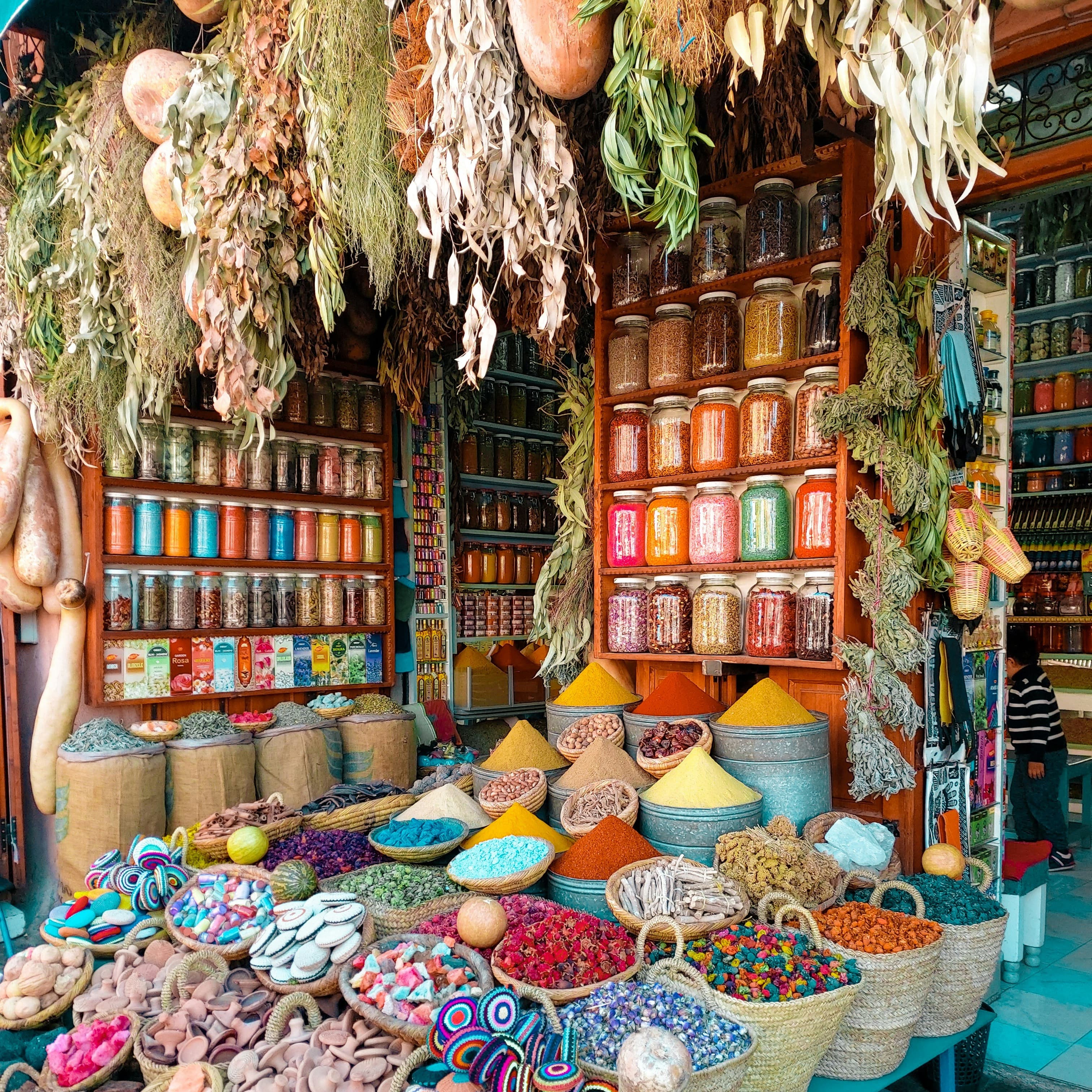 A photo of a stall at a spice market. Multicolored herbs and spices are mounded into baskets and bins on the table, a wall contains jars of multicolored beans and spices, and more herbs are hanging from the roof of the stall.