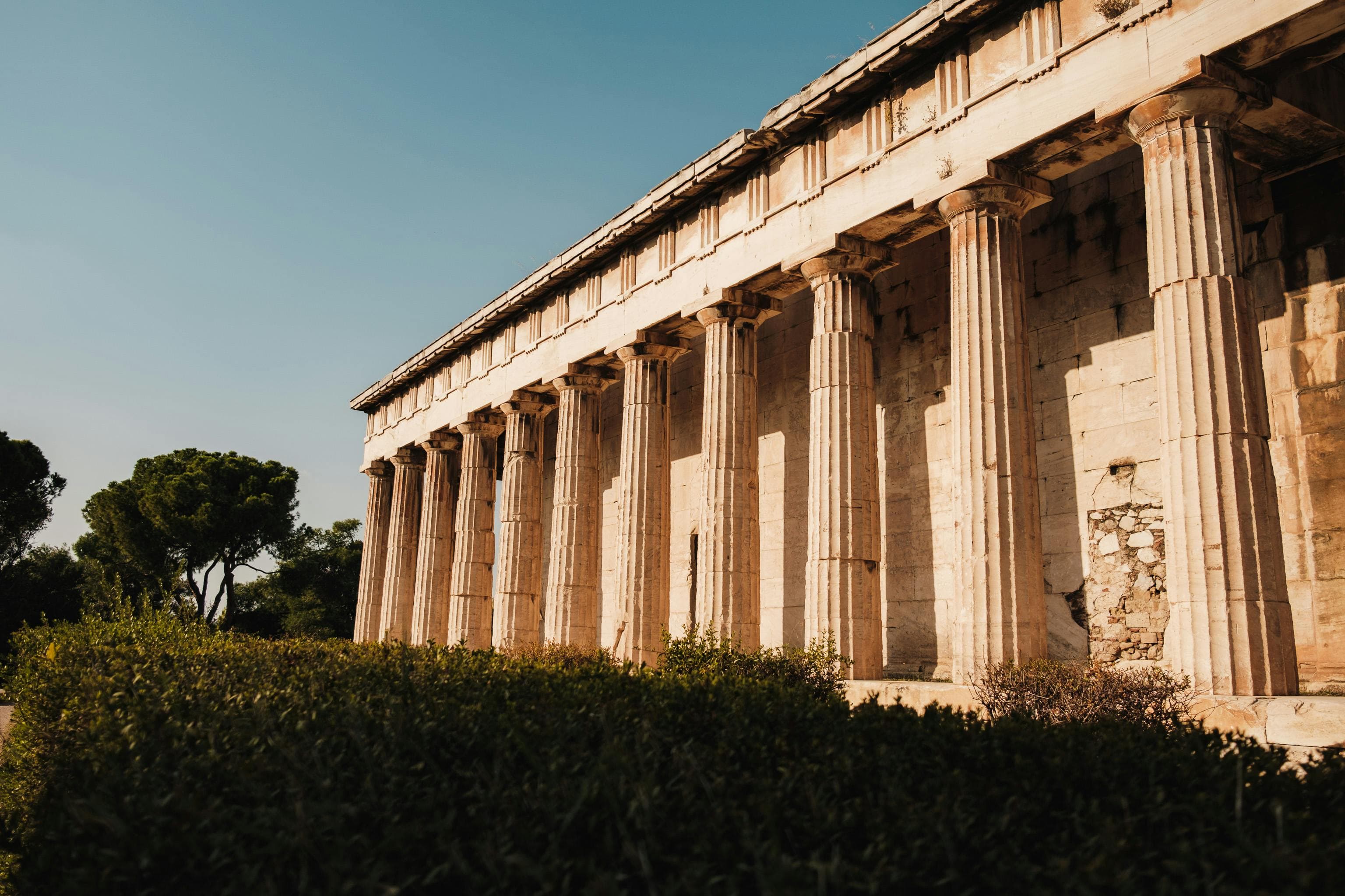 An Ancient Greek public building with large stone columns stands before a garden.