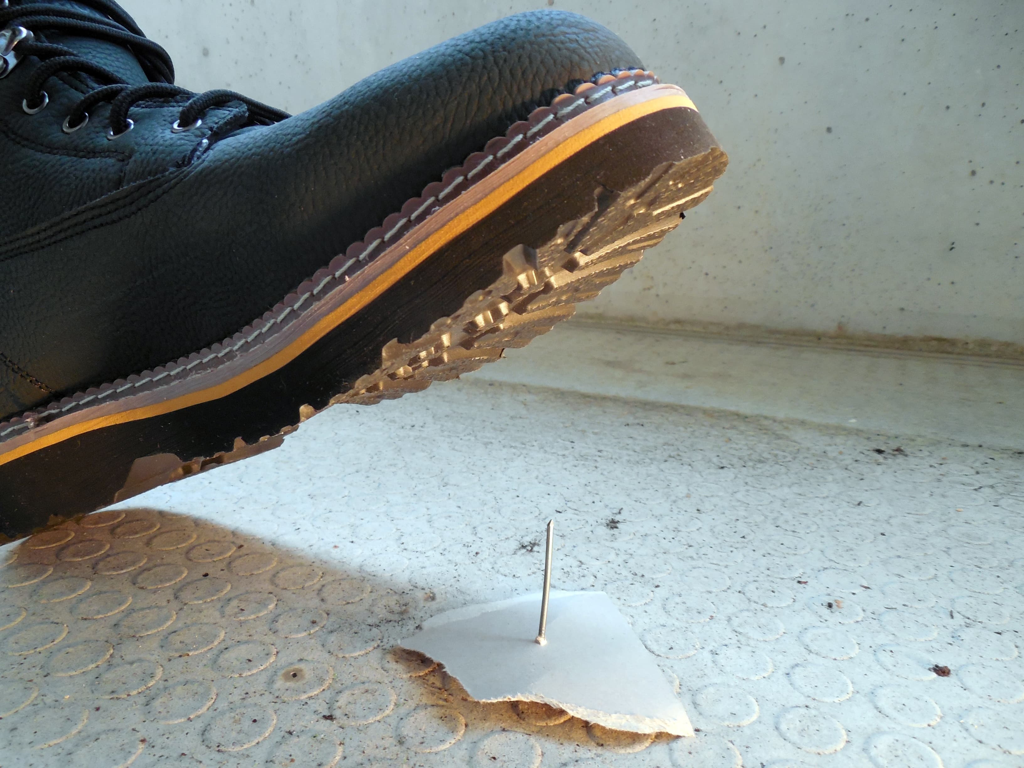 A photo of a heavy boot in the middle of taking a step. A long sharp nail is sticking up from the ground, and clearly the person in the boot is about to step on it.