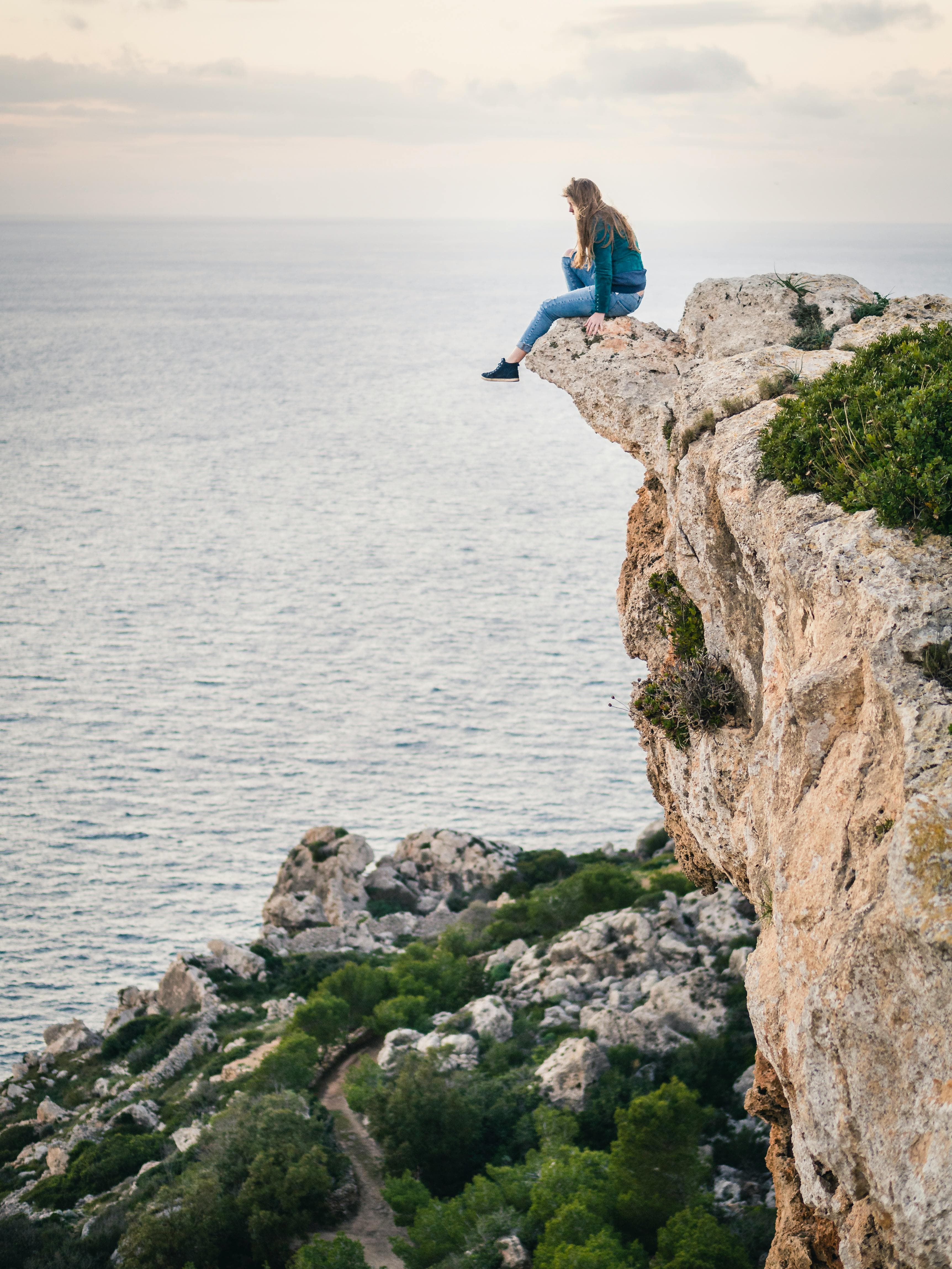 A photo of a young woman sitting with her legs dangling over the edge of a cliff by the sea at sunset.