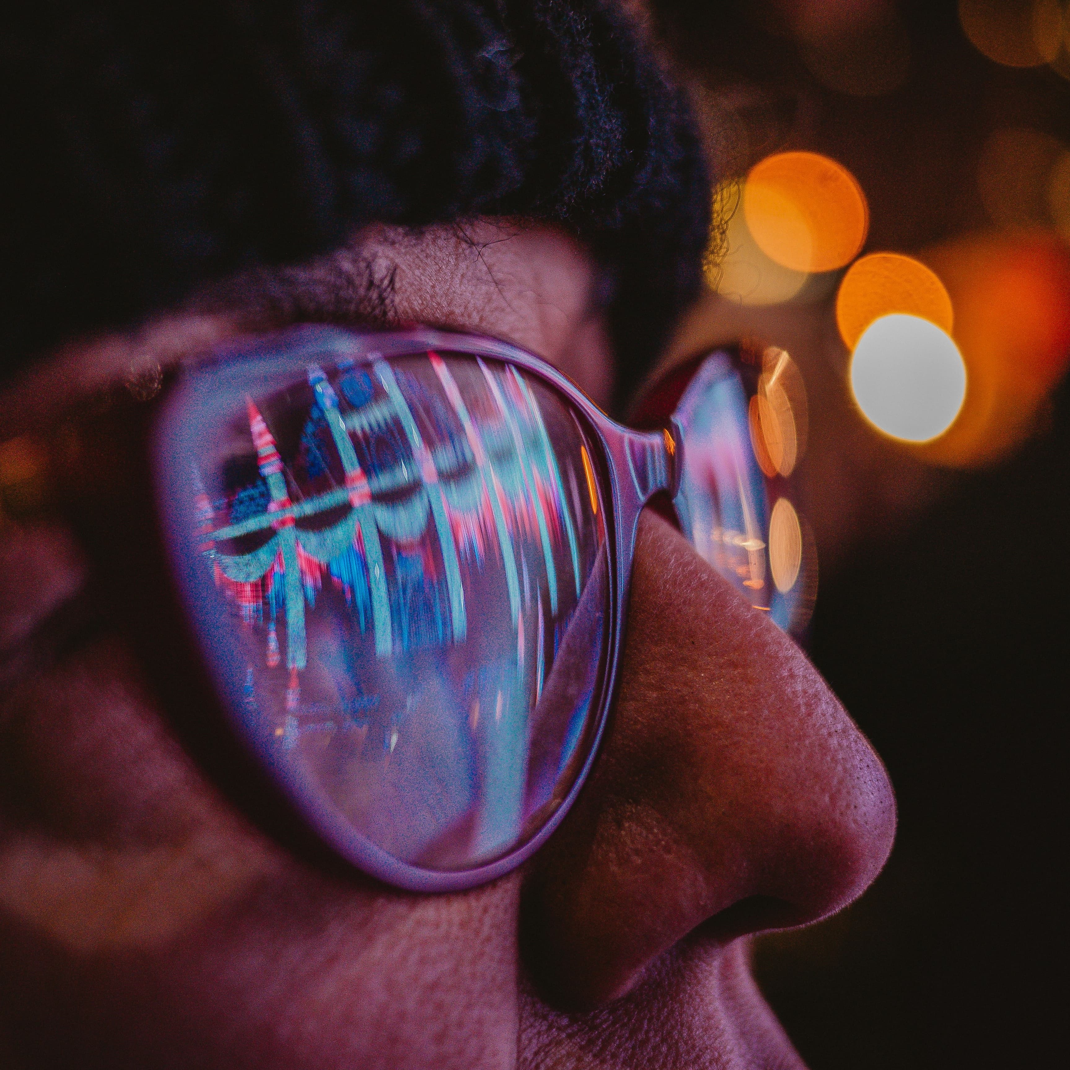 An up-close photo of someone wearing glasses. It is dark and we can see lights reflected in the panes of the glasses.