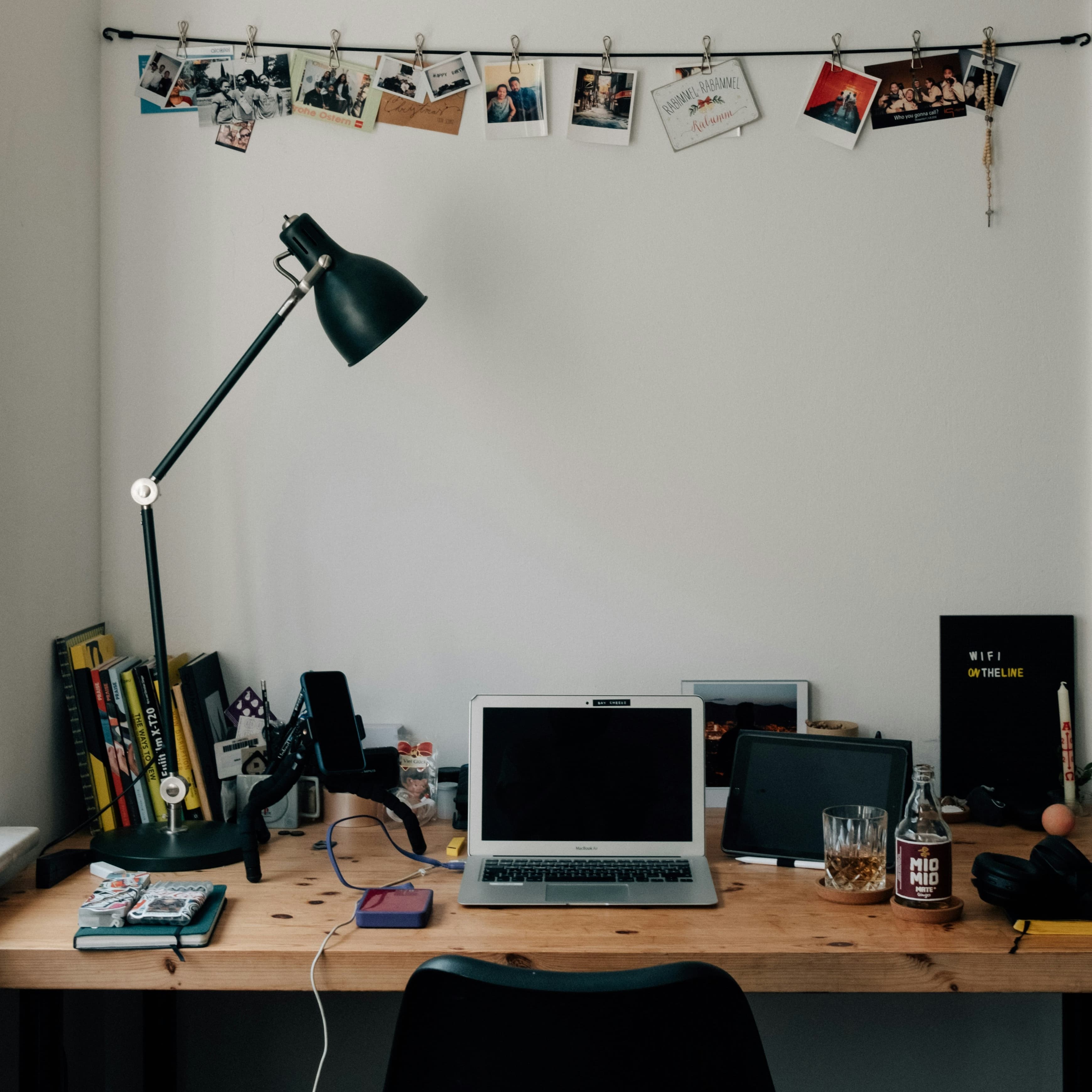 A photograph of a crowded wooden desk against a white wall. A silver laptop sits in the middle, but there is also a phone, an tablet, some drinks on coasters, a stack of books, a phone stand, a notebook, a large black task lamp, and some other clutter. On the wall, some photographs are pinned to a string.