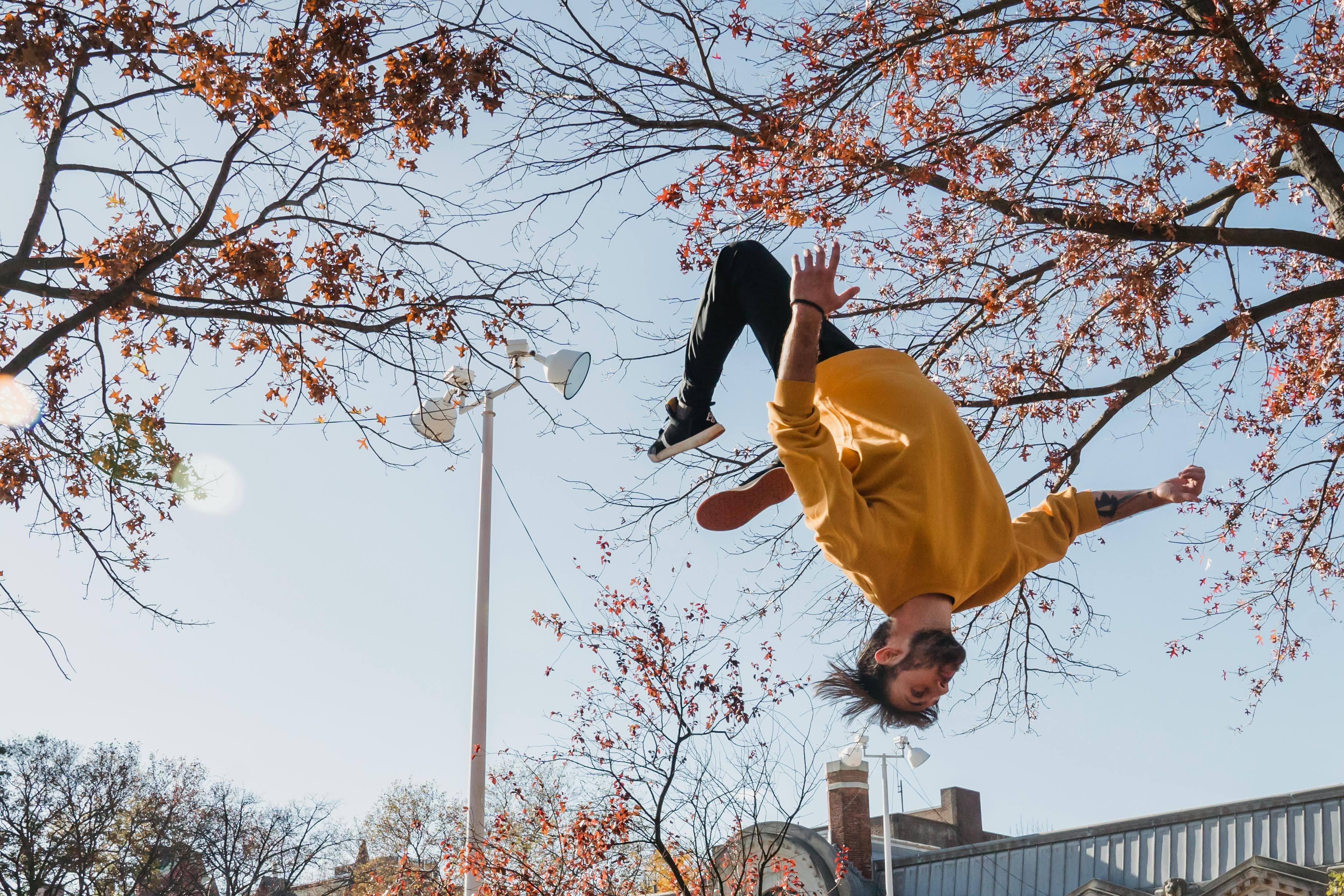 A photo of a man in a yellow sweatshirt in the middle of doing a hands-free backflip on an autumn day.