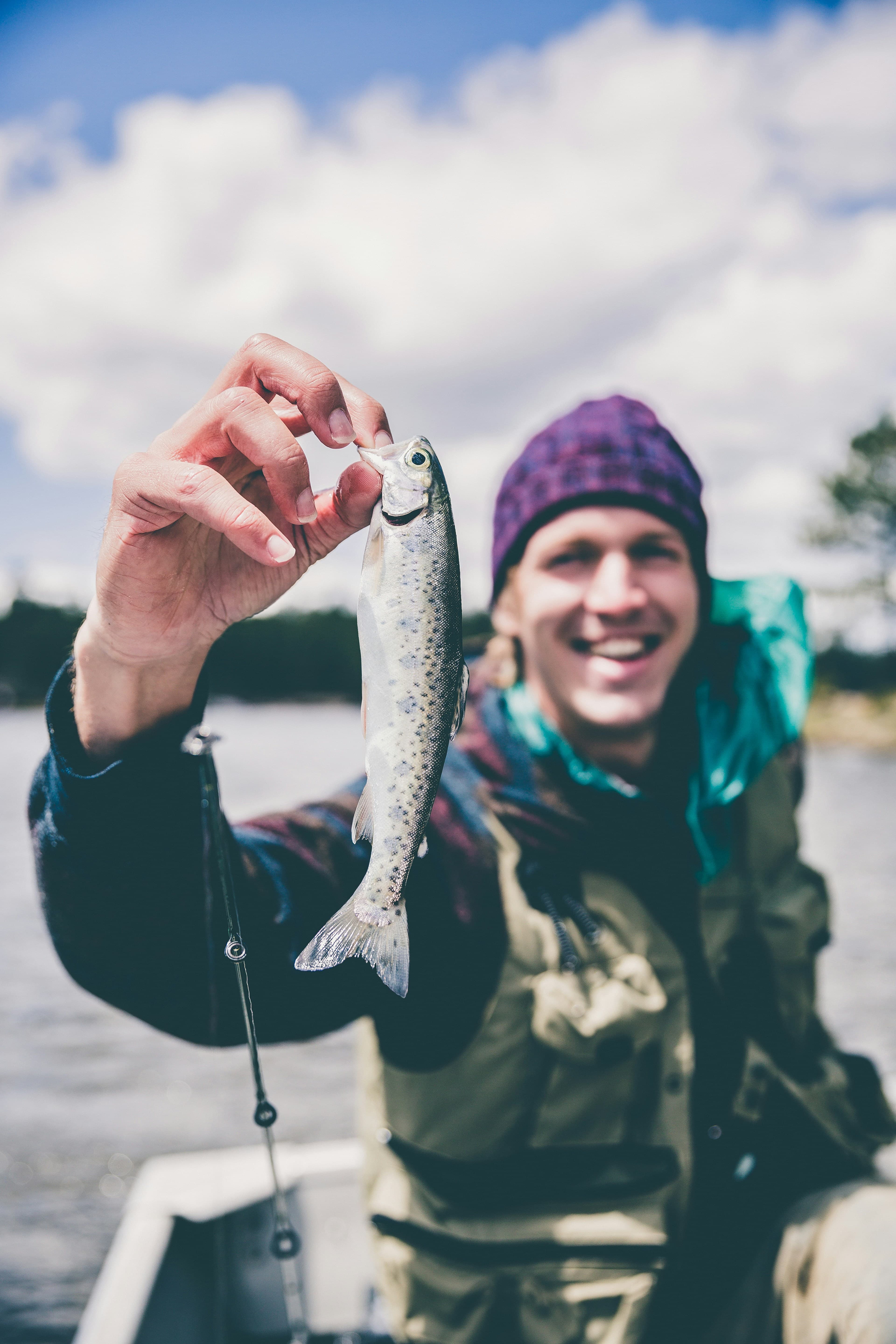 A man in a small fishing boat hold a small fish up to the camera. It is about 6 inches long.