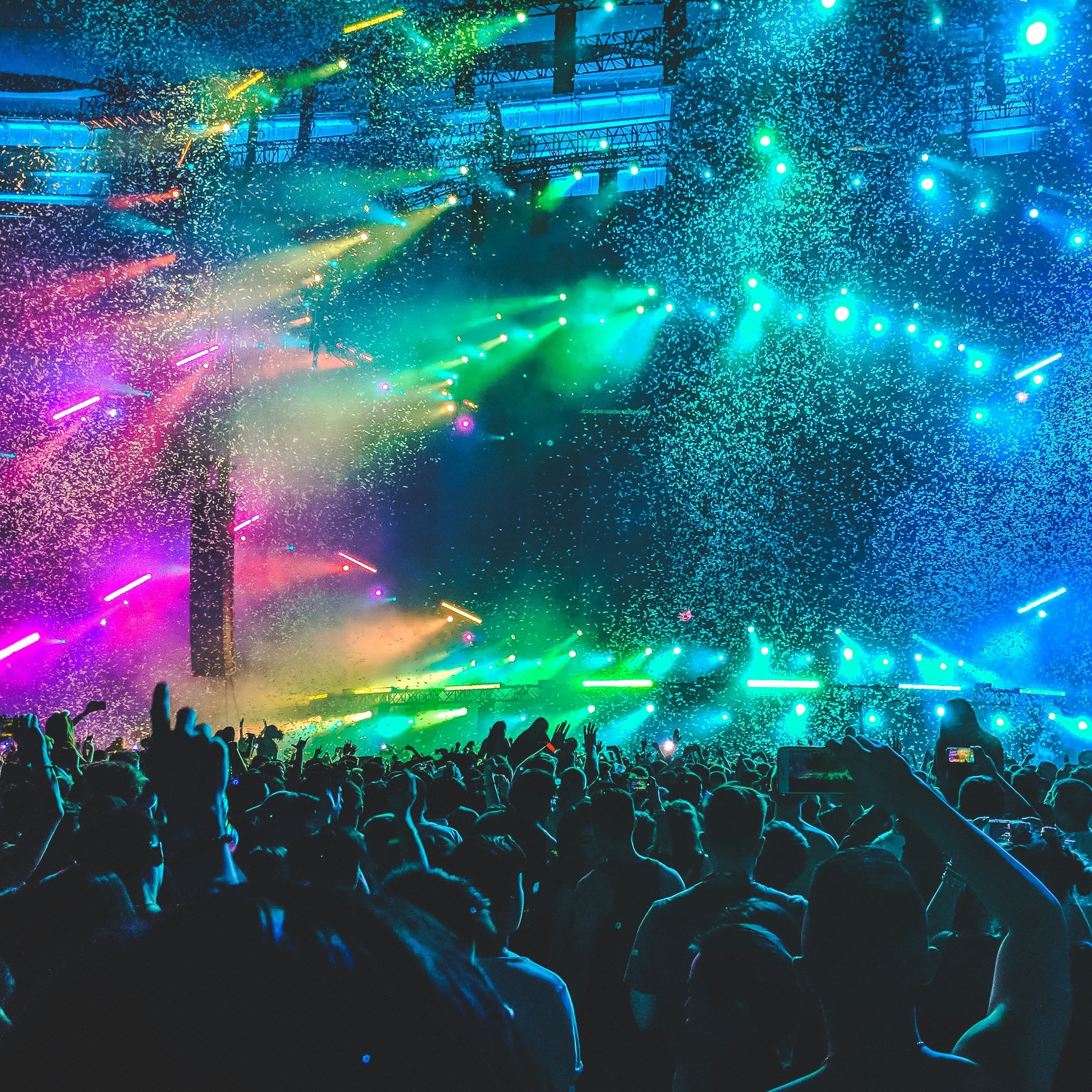 A crowded concert. It is dark, but there are rainbow stage lights. People are holding up their phones to record the stage.