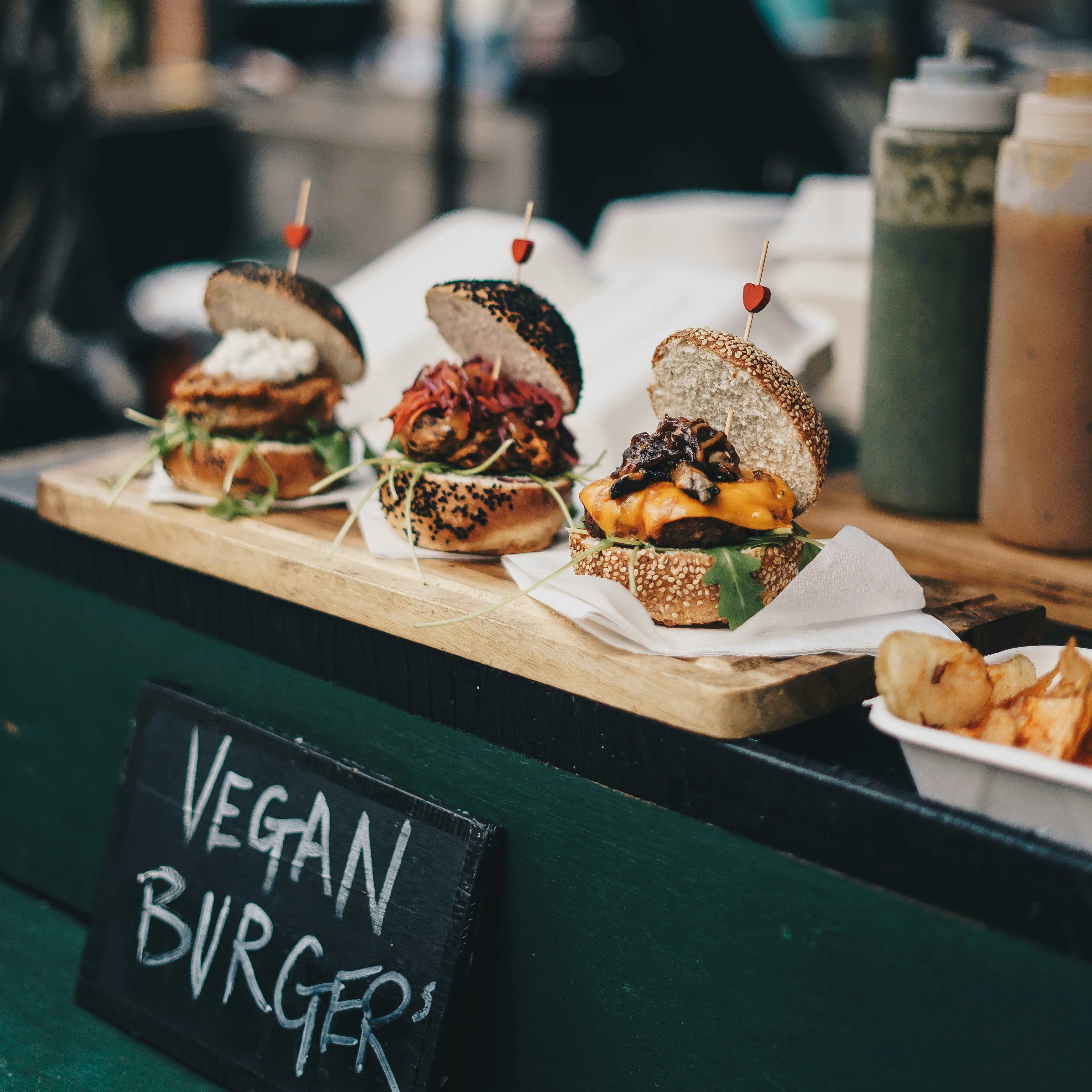 A photo of three artisanal burgers lined up on a cutting board. The buns and contents of the burgers all look a little different. Lined up behind them we can see several bottles of sauce, and a sign black handwritten sign with white writing sits beneath them, reading "Vegan Burgers."