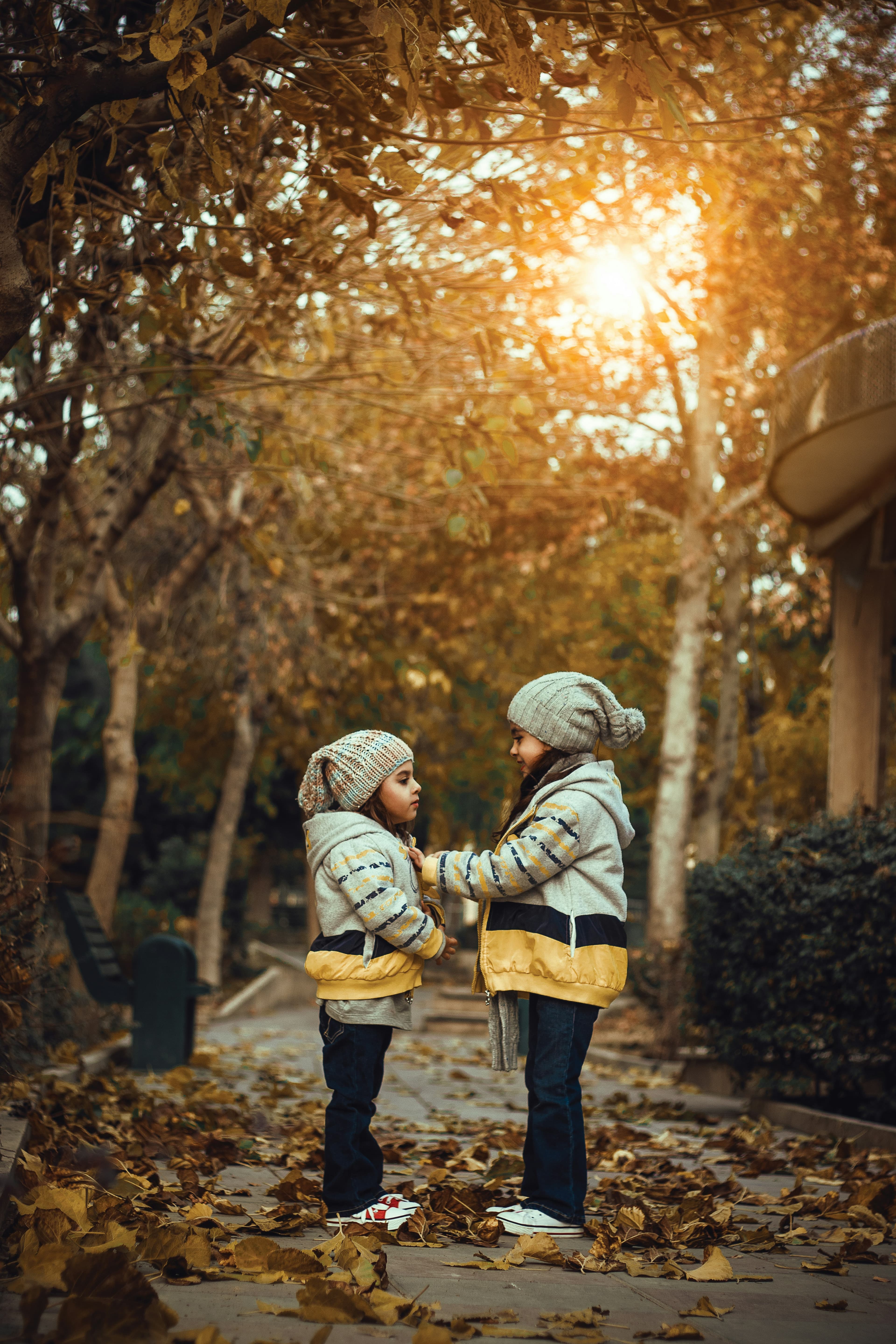 Two young girls stand outside in an autumn sunset. They are clearly sisters and are dressed in matching gray winter hats and grey and yellow jackets. They are facing each other while the older girl adjusts the younger girl's clothing.
