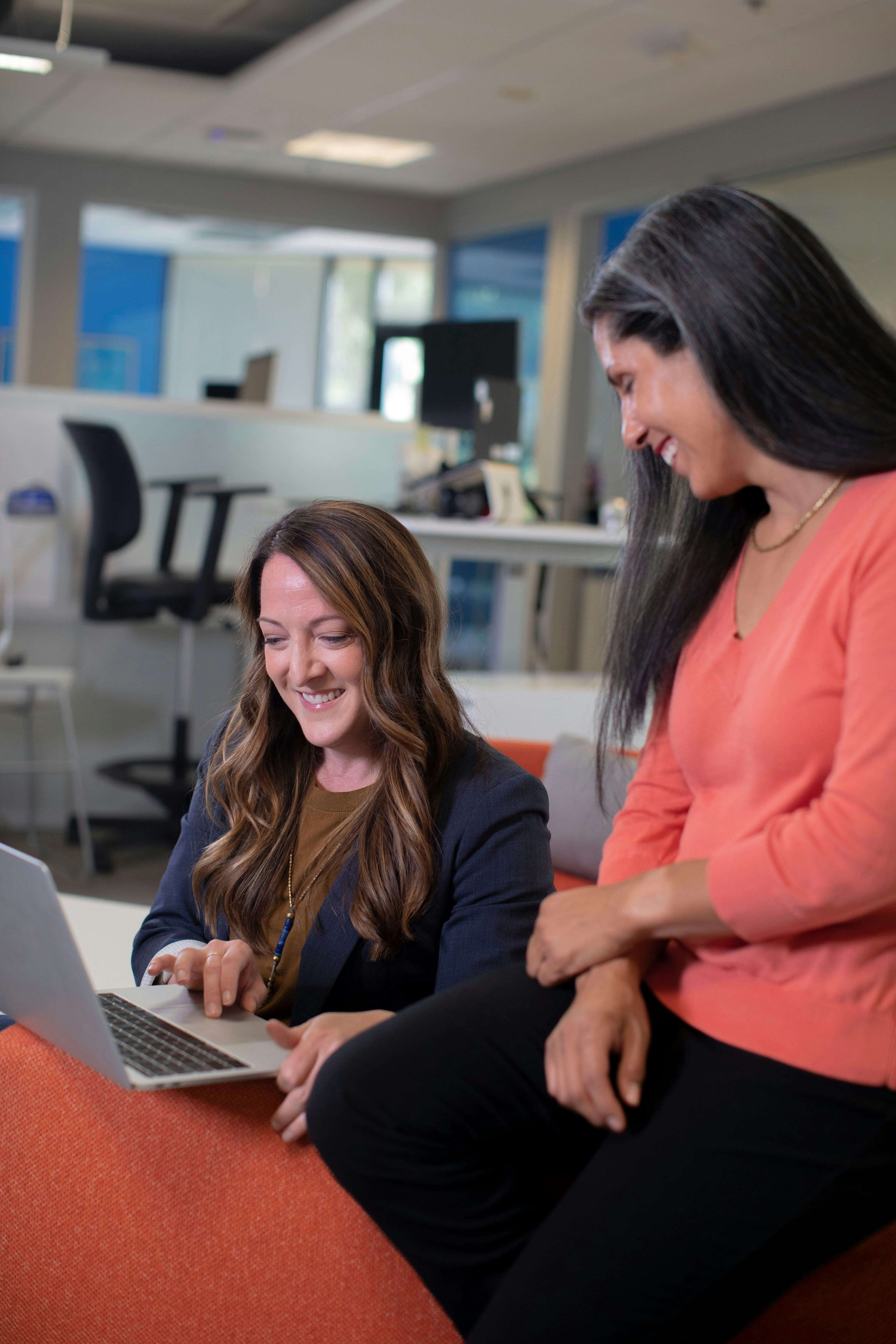 Two women with long hair in business casual are looking at a Macbook in an open plan office.