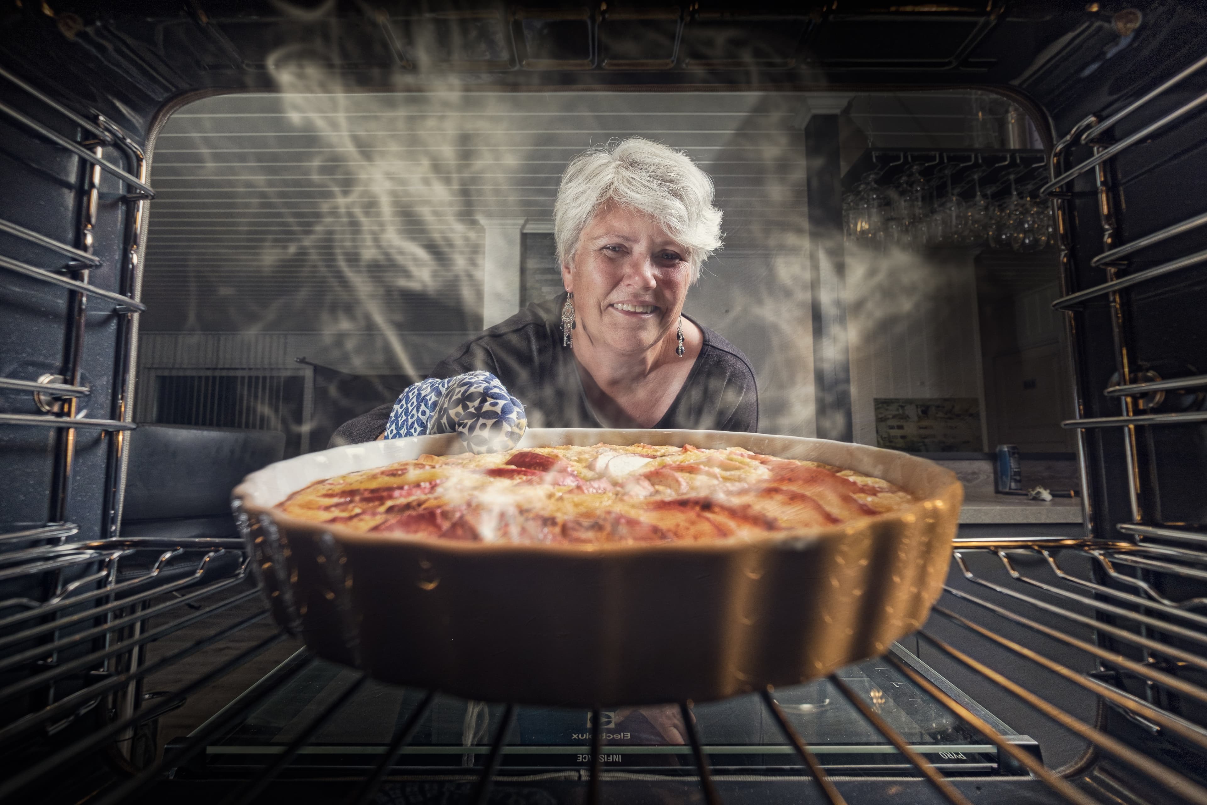 A photograph taken from inside an oven. A cake is steaming inside and an older white woman with short white hair and dangly earrings is reaching in to pull it out.