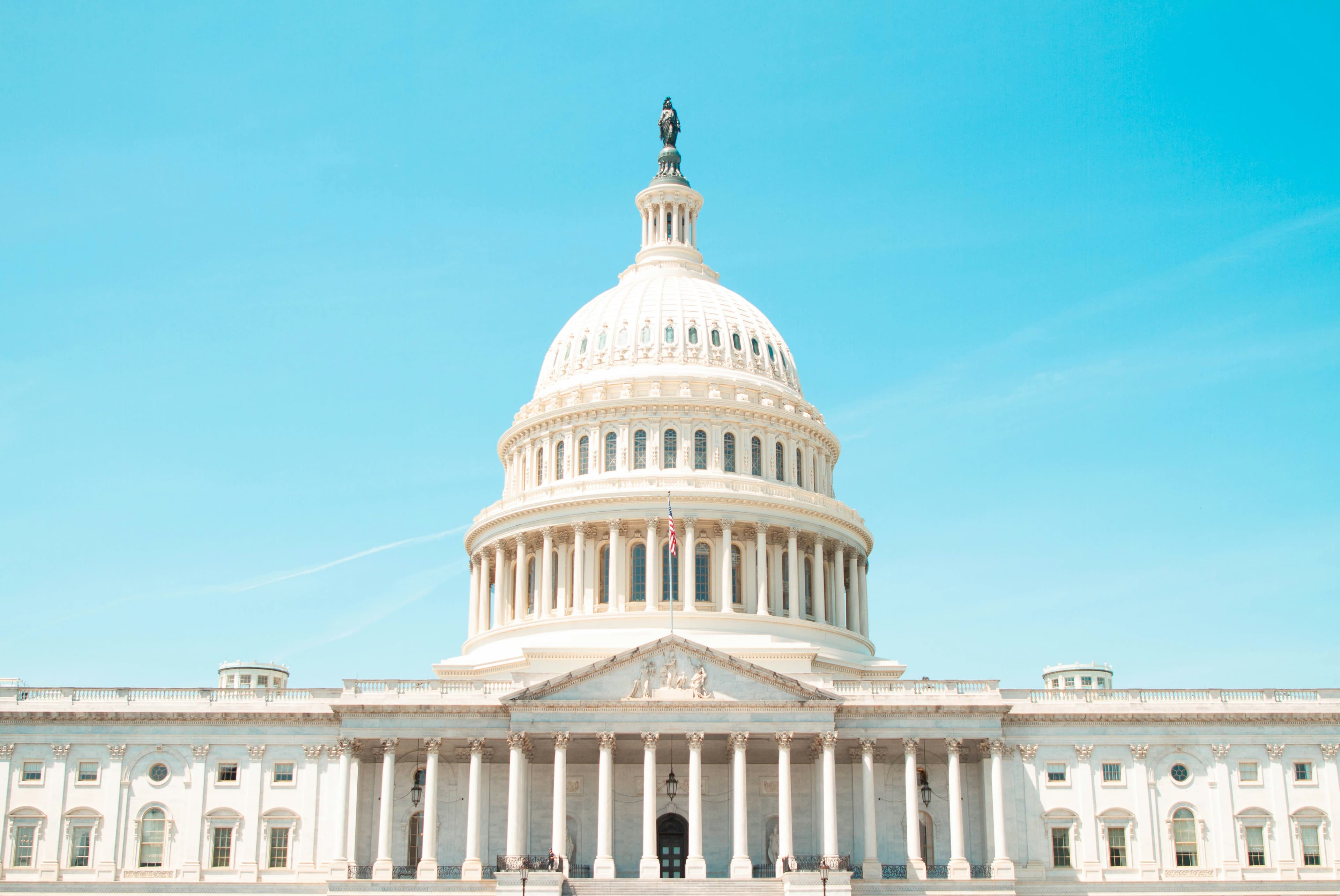 A photo of the US Capitol Building (white with columns and a large dome) on a sunny day.