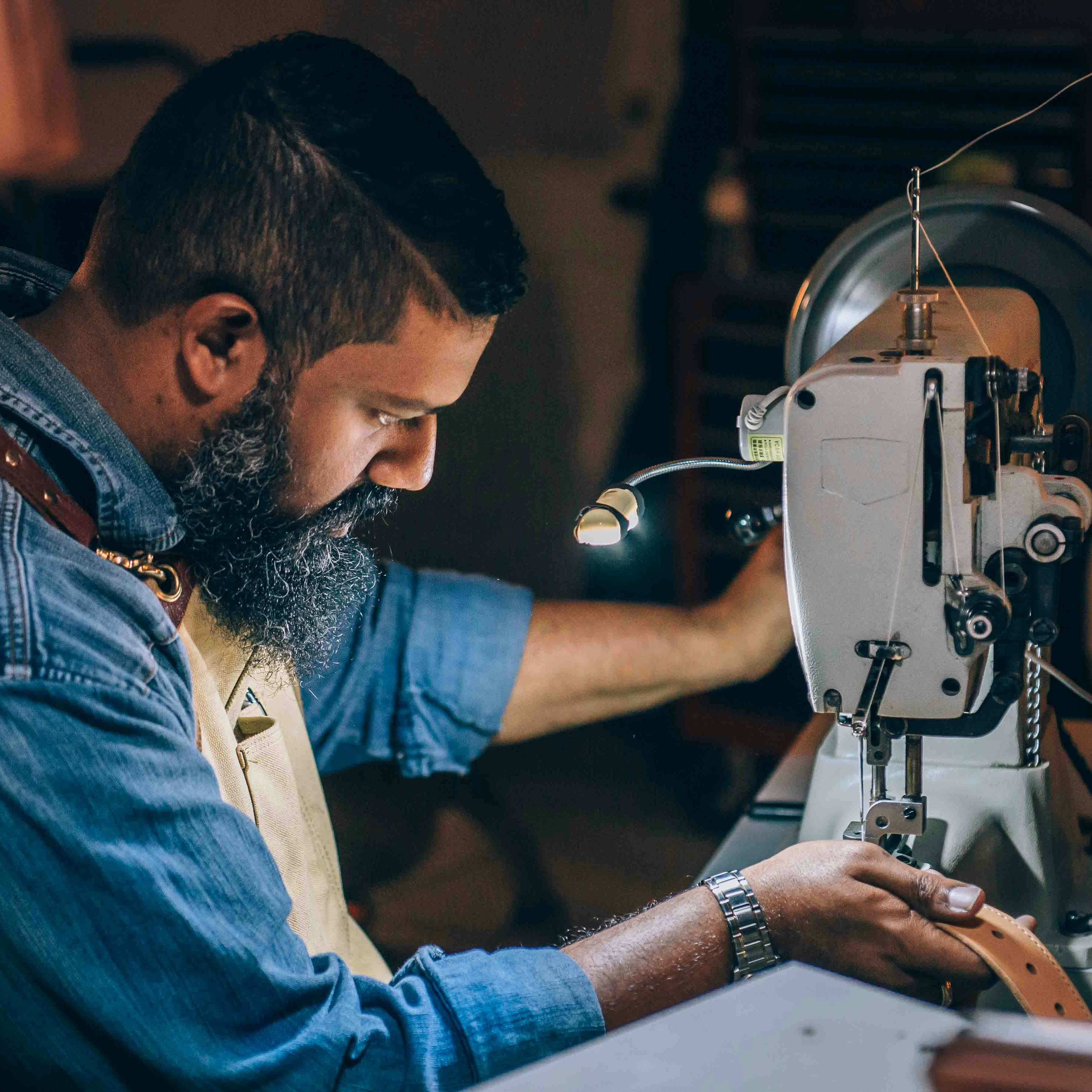 A man with a beard is wearing work clothes and is using a power tool to sew a seam into a leather belt.