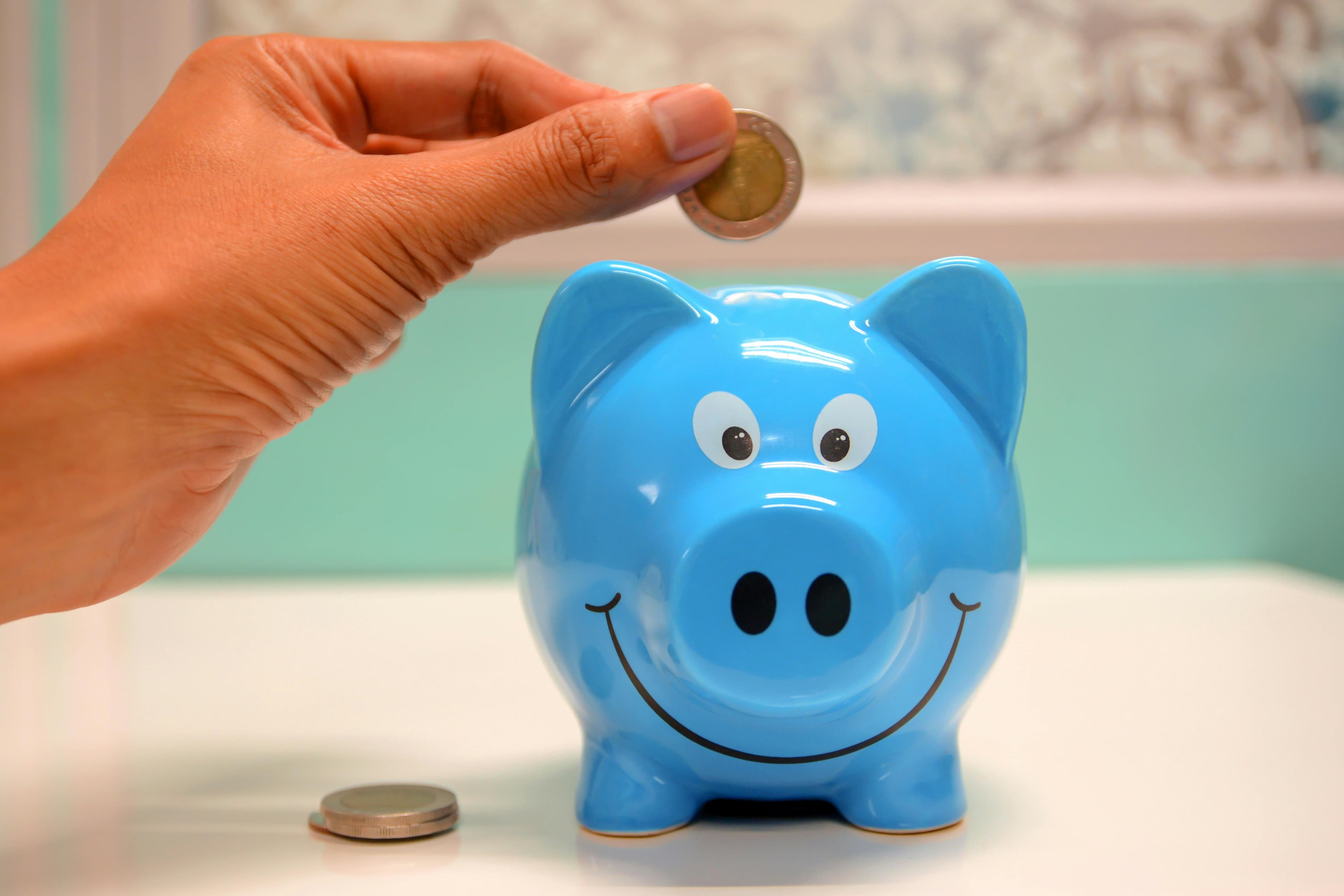 A hand is placing a coin into a smiling blue piggy bank.