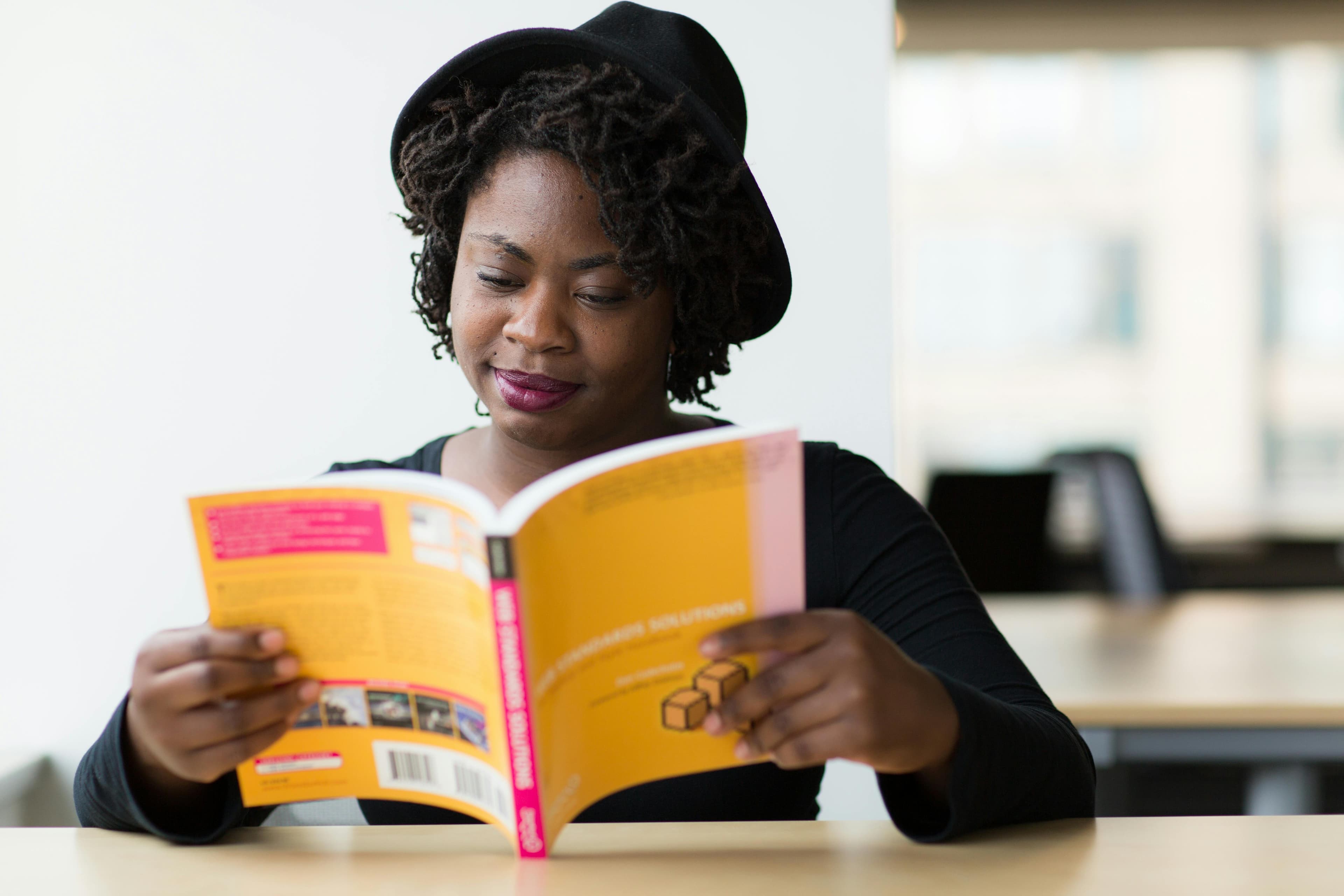 A photo of a black woman in a fedora reading a yellow book at a desk.