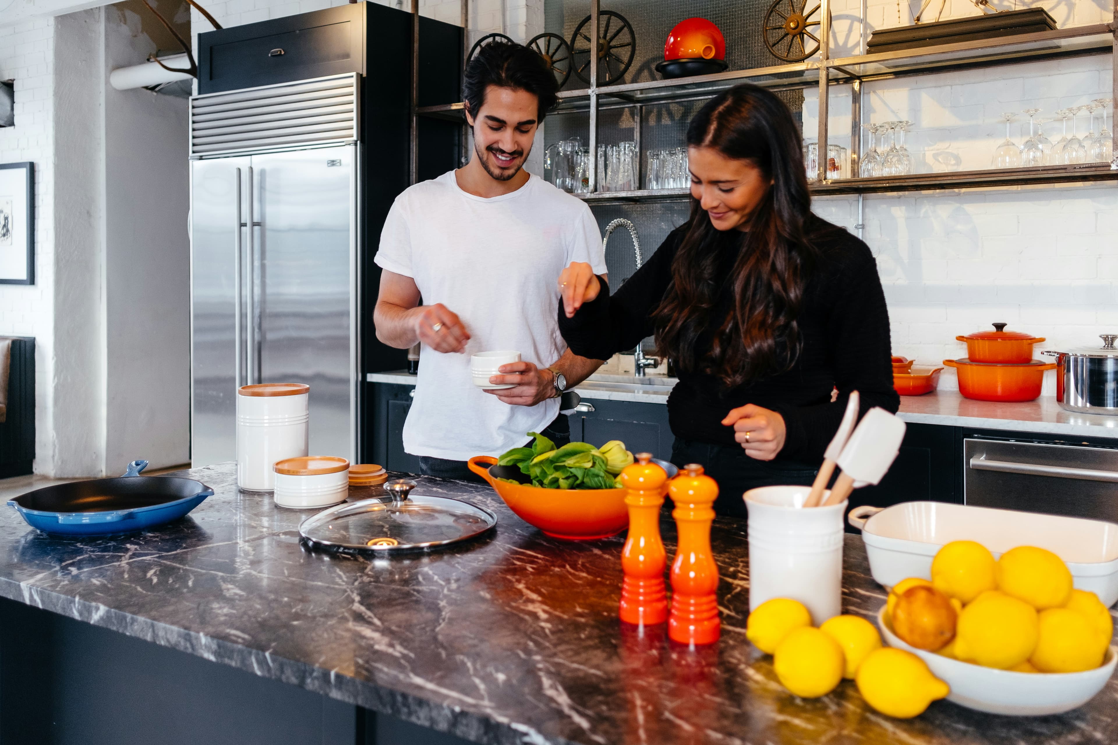 A young man and woman with dark hair cook in a fancy modern kitchen. They are smiling as they work to add the final spices and ingredients to a salad.