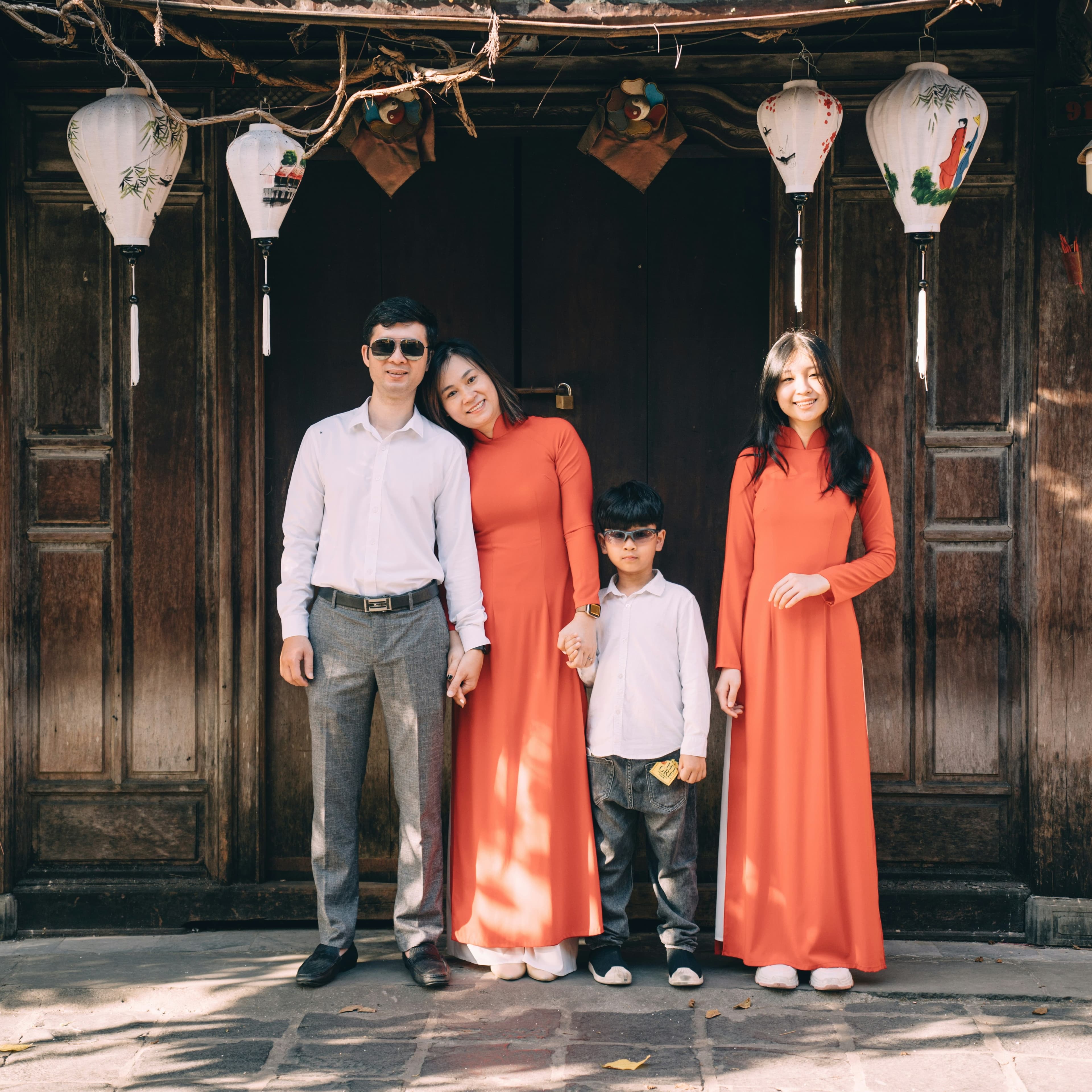 An East Asian family poses for a photo. The man and boy are in white collared shirts and gray pants, the woman and girl are in matching coral long-sleeved maxi dresses. The man woman and girl are all about the same height, and the boy seems quite a bit younger. They are posed in a historical-looking wooden doorway in an East Asian style, that is opening out into the courtyard, and several painted paper lanterns are hung above it.