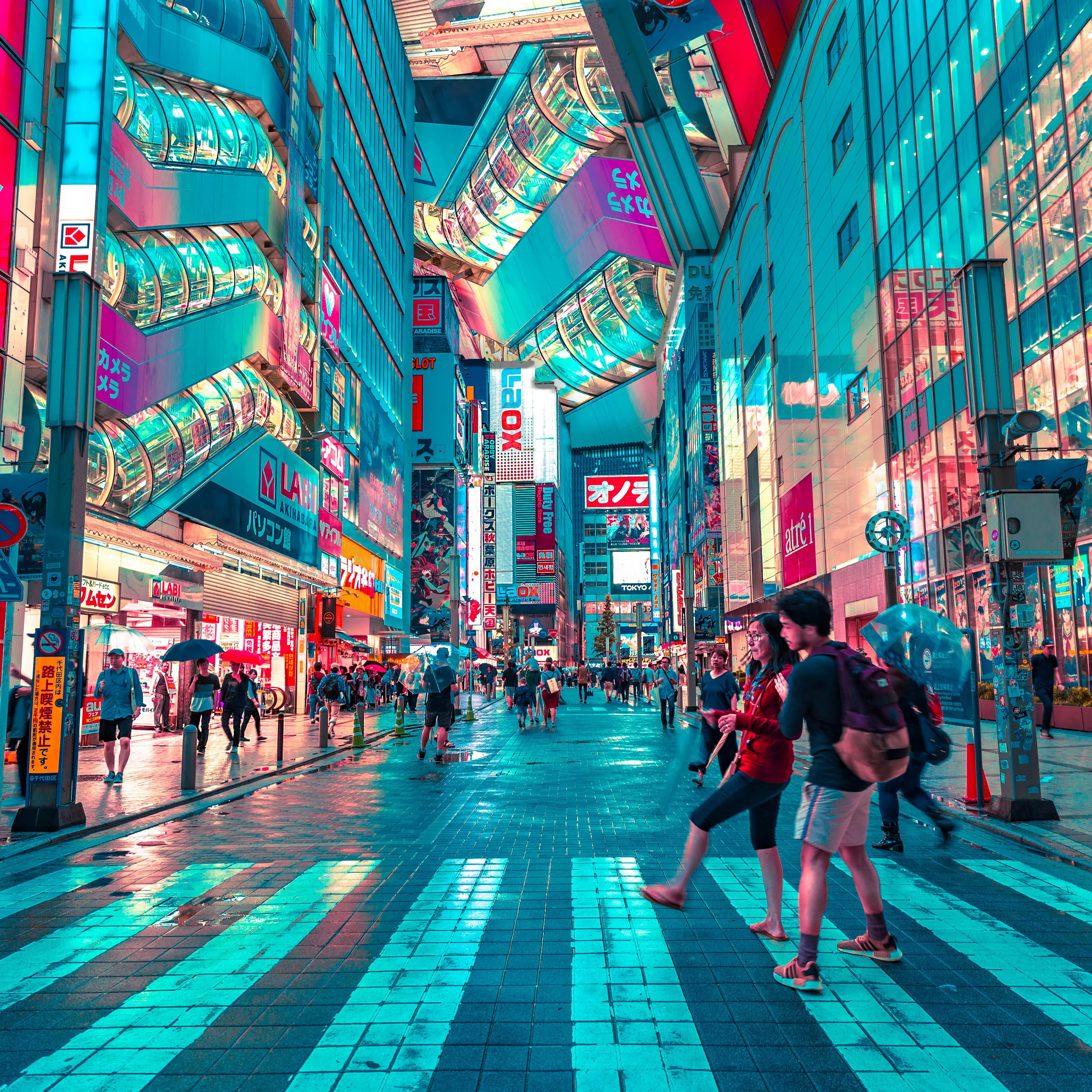 A modern city street in Asia at night. The buildings have several glass-walled walkways or large walls of glass, and the street is lit in various colors of lights (aqua, purple, red). The street is reasonably crowded with pedestrians and directly in front of us a crosswalk cuts across the road. Though there is signage for cars, the street seems to be mostly used by pedestrians.