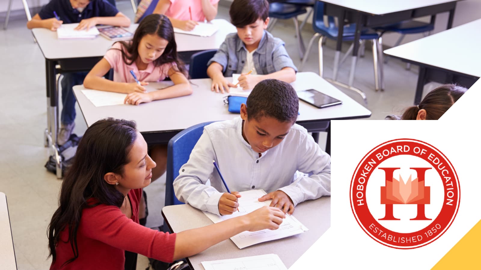 Students writing at their desks with a teacher alongside them, beside the Hoboken Board of Education seal