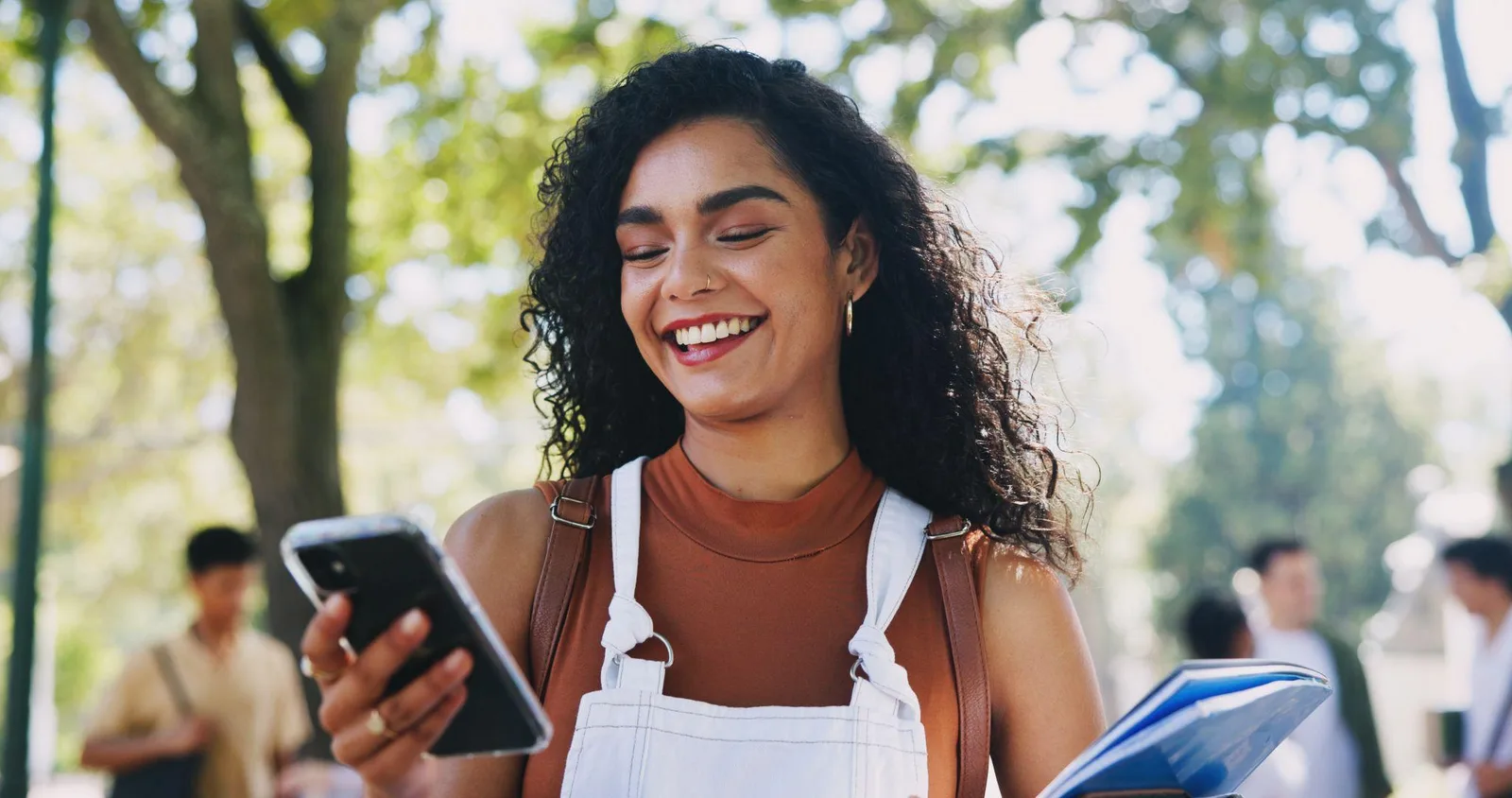 A student smiling at her phone while walking through a park, a notebook under her arm.