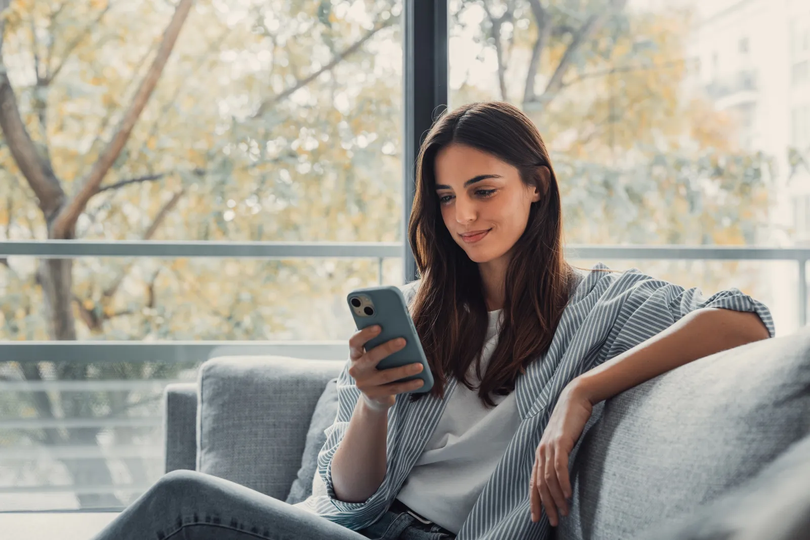 A woman relaxing on a sofa by a window, reading something on her phone.