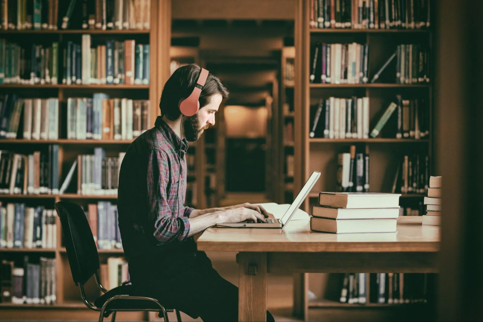 A reader with headphones on typing at a laptop at a library table, a stack of books beside him and full shelves behind.