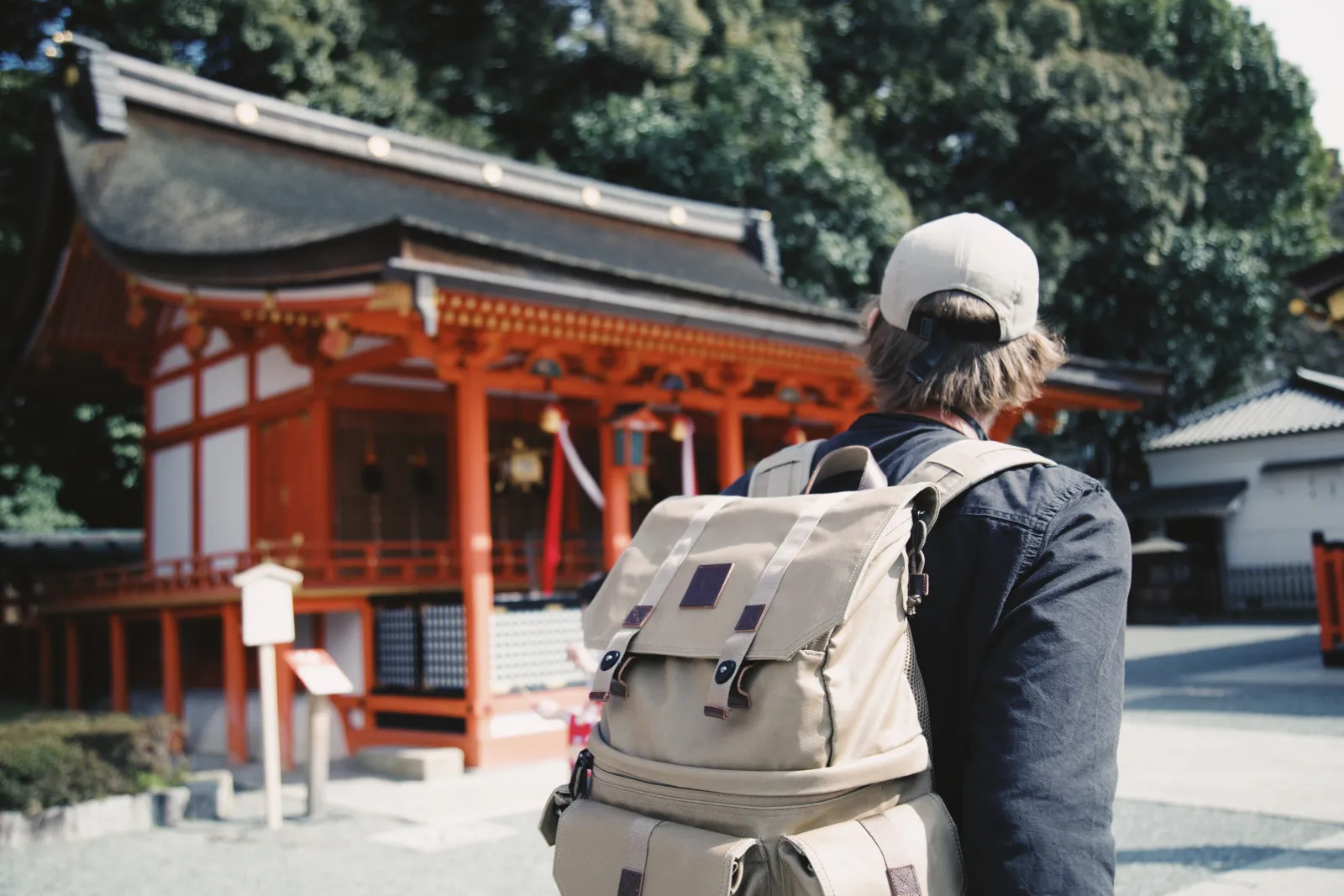A traveller seen from behind with a large backpack, facing the vermilion gate of a Japanese shrine.