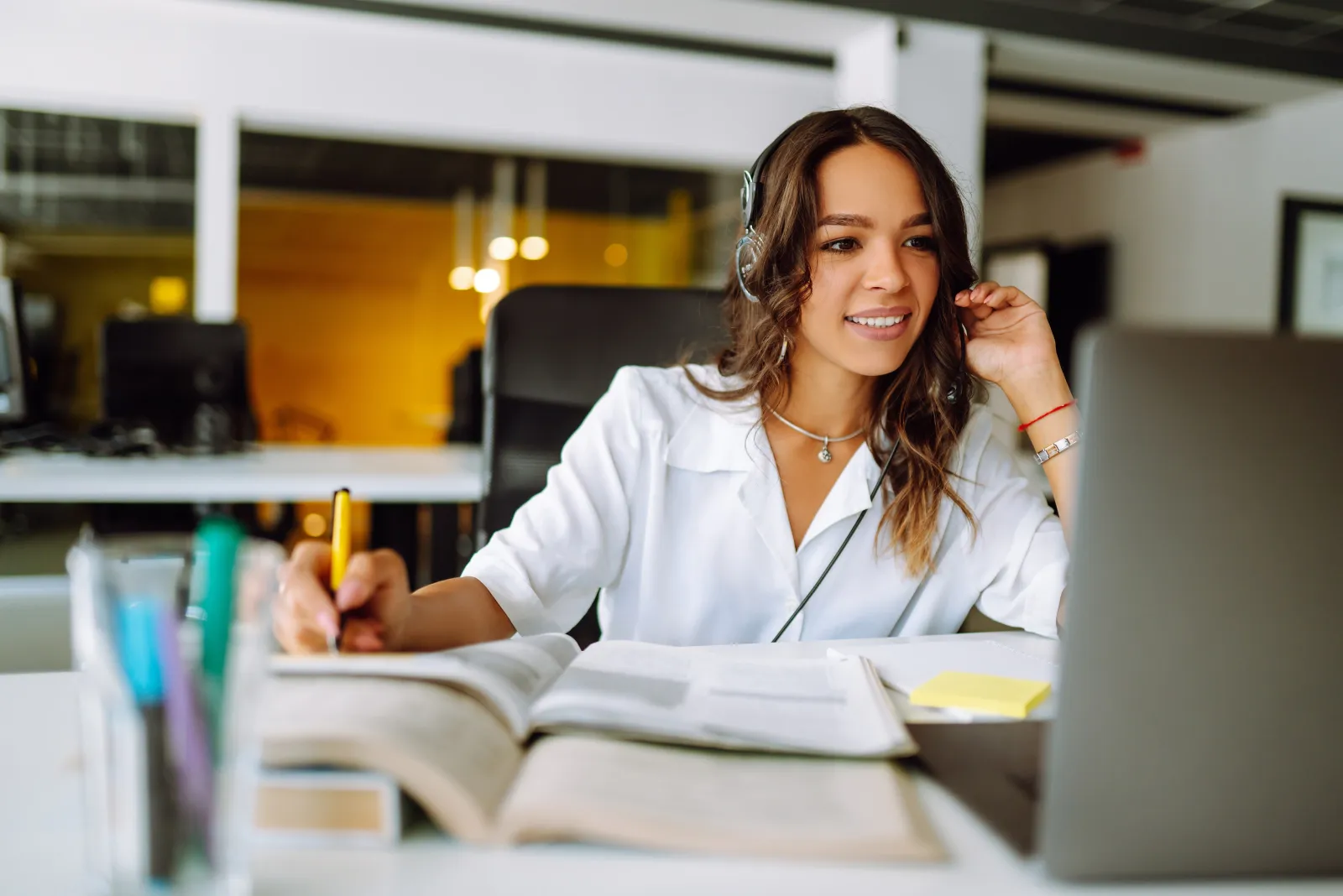 A support agent in a headset smiling at her screen, taking notes in an open ledger.